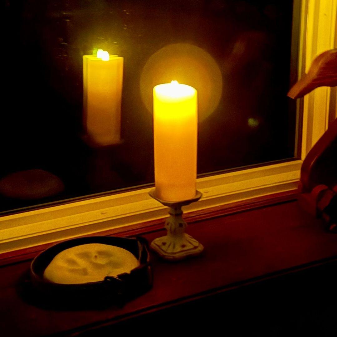 A lit LED candle on a windowsill next to a dog collar, reflected in the window
